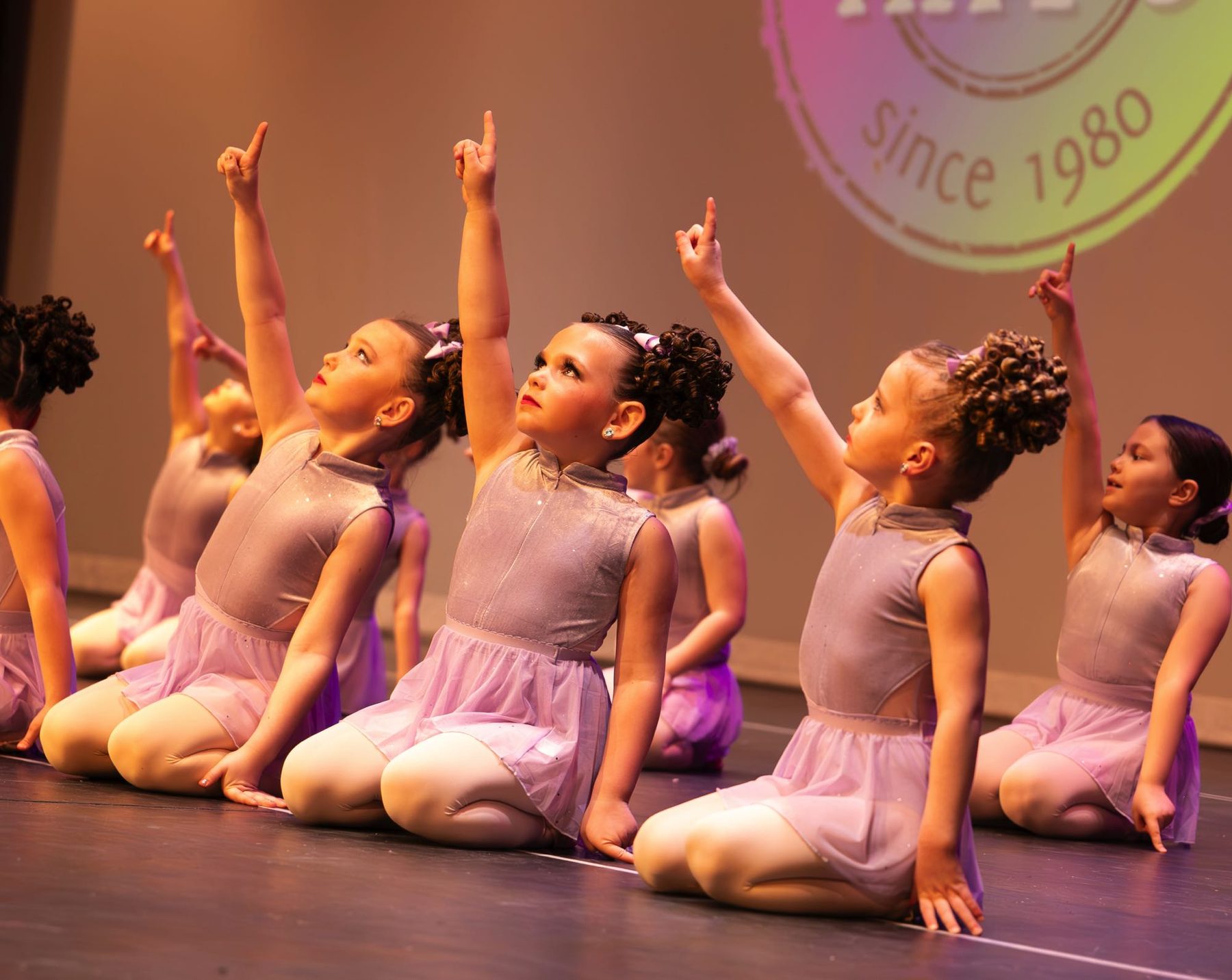 Young Kay's Act II dancers kneeling on stage, each pointing skyward, with the Kay's 'since 1980' logo projected behind them